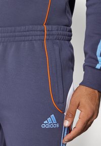 Pantalones jogger en color navy, confeccionados con tela suave, con ribetes naranjas, bolsillos laterales y un logo bordado en azul claro. Cintura elástica texturizada.