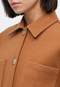 Ruskea villasekoitekankainen paitatakki, jossa on terävä kaulus, kaksi etutaskua ja suuret, sileäpintaiset napit. Pehmeä koostumus.