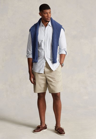 Šviesiai mėlyna dryžuota marškiniai su sagomis, smėlio spalvos šortai, tamsiai mėlynas pynuotas megztinis užmestas per pečius, rudi laivų batai su raišteliais.