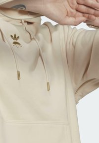 Krämfärgad hoodie i mjukt tyg med dragkedja framtill och en guldfärgad logga. Texturerade detaljer på ärmsluten och dragsko.