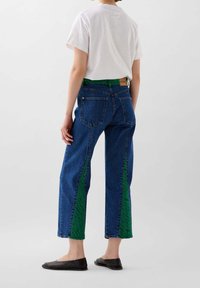 Jeans de mezclilla azul de cintura alta con rayas verdes verticales; combinados con una camiseta blanca y flats negros, mostrando un ajuste casual y moderno.