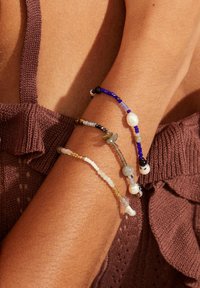 Drei Perlenarmbänder in verschiedenen Farben, darunter Blau, Weiß und Gold, bestehen aus unterschiedlichen Materialien und Größen und liegen auf einem gebräunten Arm.