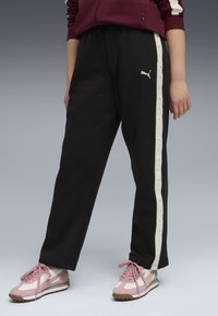 Pantaloni da tuta neri con una striscia laterale bianca che presenta un piccolo logo. Abbinati a sneakers rosa chiaro, con accenti bianchi a contrasto e suole marroni.