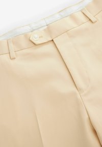 Beige Hochbundhosen mit Knopfverschluss, Gürtelschlaufen und sichtbaren Nähten auf glattem Stoff.