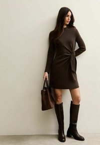Brązowa sukienka z długim rękawem z wiązaniem w talii, zestawiona z brązowymi skórzanymi kozakami do kolan i dopasowaną torebką z fakturą.