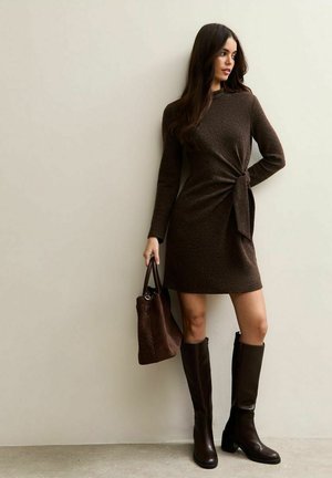 Vestito marrone a maniche lunghe con dettaglio a nodo in vita, abbinato a stivali di pelle marroni al ginocchio e una borsa coordinata in tessuto lavorato.