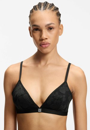 Junge Person mit geflochtenem Haar trägt ein schwarzes florales Spitzen-Bralette mit dünnen Trägern und "HUGO"-Logo am Bund.