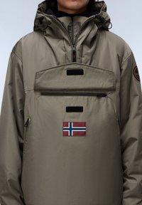Persoon in een olijfgroene jas met een voorzak met rits, een Noorse vlag patch en een verstelbare capuchon met trekkoorden.