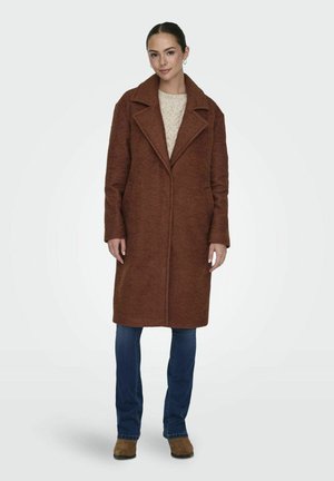 Manteau en laine marron avec un col en V, une ouverture sur le devant et des poches latérales. Porté sur un pull léger en tricot et un jean bleu. Matériau doux et texturé.