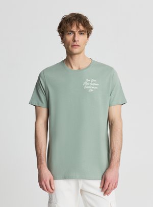 Giovane uomo con capelli ricci che indossa una maglietta verde chiaro con piccolo testo bianco sul petto e pantaloni bianchi su sfondo neutro.