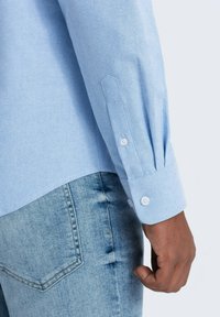 Camisa de botón de manga larga azul claro con puños, tela texturizada, brillo sutil, combinada con jeans de mezclilla de lavado claro con bolsillo trasero.