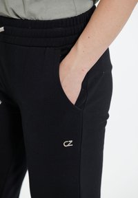 Svarta joggerbyxor med elastisk midja, sidofickor och en liten silverfärgad logotypdetalj på vänster underben. Slät tygtextur.