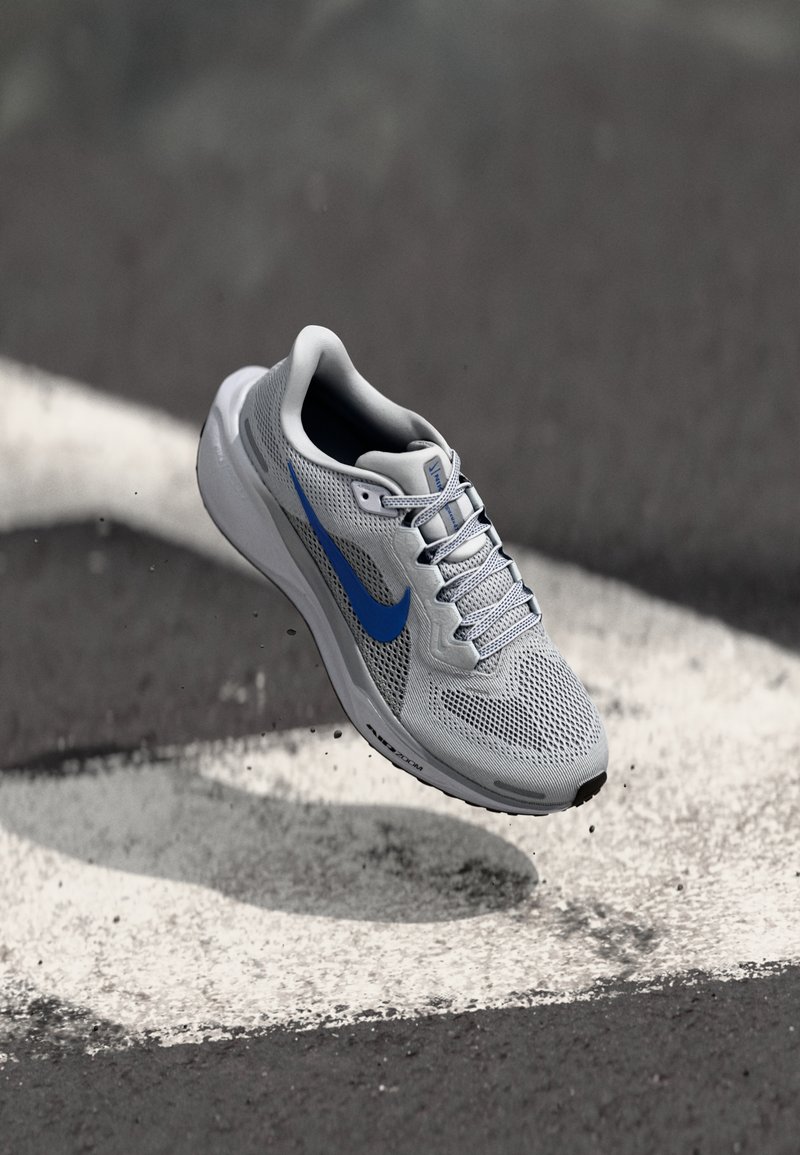 Adidași sport de culoare gri deschis, cu un superior din plasă respirabilă, swoosh-ul albastru Nike și o talpă texturată, suspendați deasupra unei suprafețe de asfalt.