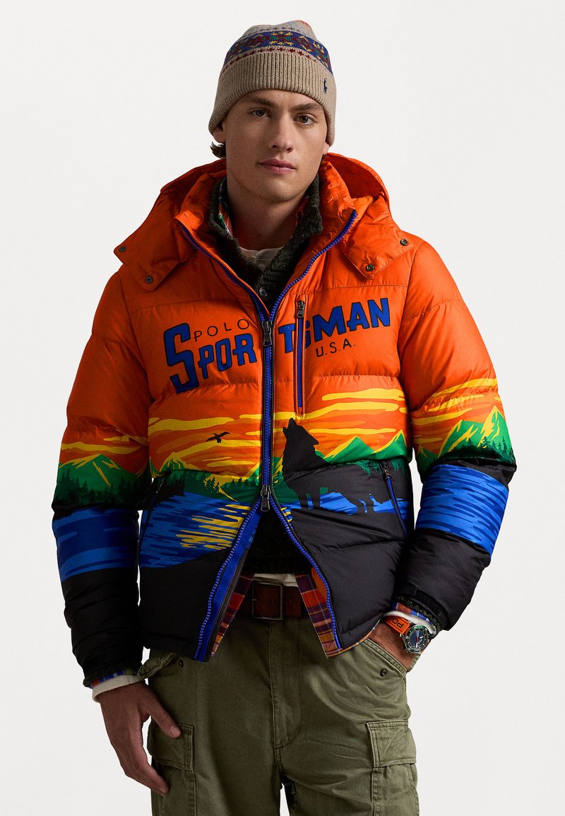 Bunte Pufferjacke aus orangefarbener Stoff mit Berg- und Landschaftsgraphic, blauen Akzenten und einem Reißverschluss vorne. Kombiniert mit Cargo-Hosen.