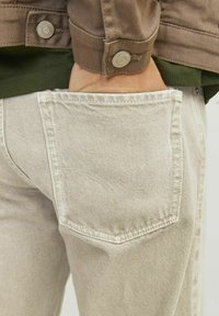 Jeans de color beige claro con un detalle de bolsillo trasero que presenta una costura sutil. La cinturilla muestra una tela verde superpuesta debajo de una chaqueta marrón.