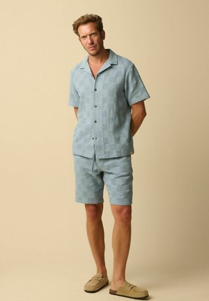 Man draagt lichtblauw gestructureerd kort mouw overhemd met knopen en bijpassende korte broek, beige instapschoenen, staand voor een effen beige achtergrond.