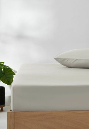 Minimalistisches Schlafzimmer mit einem Holzrahmenbett, beigem Spannbettlaken und Kissen sowie einer grünen Topfpflanze vor einer weißen Wand.