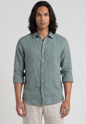 Giovane uomo dalla pelle media e con lunghi capelli scuri indossa una camicia verde abbottonata con le maniche rimboccate e pantaloni beige chiaro, in piedi in modo neutro su sfondo bianco.