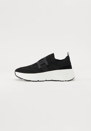 Schwarzer gestrickter Slip-on-Sneaker mit dicker weißer Sohle und elastischem Riemendetail auf weißem Hintergrund.
