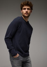 Tmavě modrý pletený svetr s texturovaným vzorem, kulatým výstřihem a dlouhými rukávy, doplněný o světle šedé džíny.