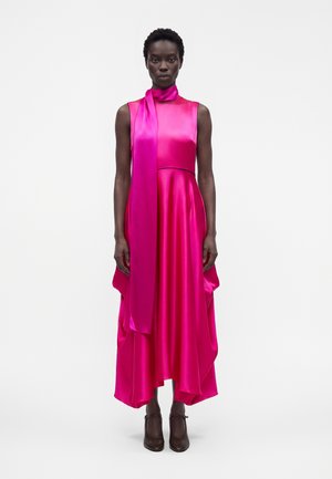 Femme portant une robe en satin rose vif sans manches avec un ourlet asymétrique et une écharpe assortie, associée à des chaussures à talons hauts marron foncé.