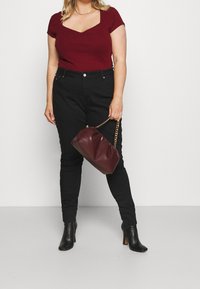 Kvinna i en figursydd bordeaux kortärmad topp, svarta högmidjade skinny jeans, svarta klackiga ankelstövlar, hållande en bordeauxväska med guldkedjesnor.