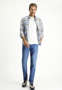 Lichtblauw en beige geruit overhemd over een wit t-shirt, gecombineerd met blauwe spijkerbroek en grijs-zwarte sneakers. Rechte pijpen en opgerolde mouwen.