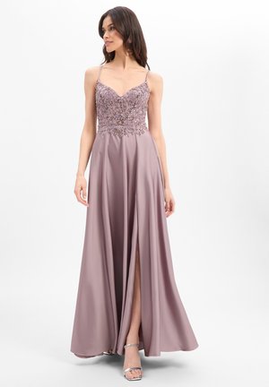 Frau in einem mauvefarbenen Spaghettiträgerkleid mit perlenverziertem Oberteil und hohem Beinschlitz, trägt silberne offene Absatzschuhe und steht vor einem weißen Hintergrund.