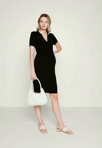 Vestido preto justo com mangas curtas e decote em V, combinado com uma bolsa branca texturizada e sandálias slip-on de cor clara.