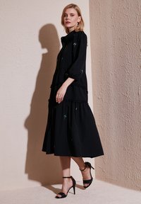 Rochie neagră cu volane, decorată cu broderie florală albastră. Are mâneci lungi, tiv ondulat și guler înalt. Pur și simplu se asortează cu tocuri negre cu curea.