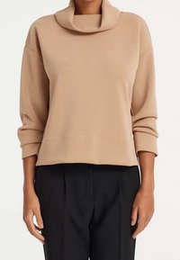 Maglione beige con collo ampio, maniche lunghe e vestibilità rilassata. Il materiale appare morbido con una texture liscia, abbinato a pantaloni neri.