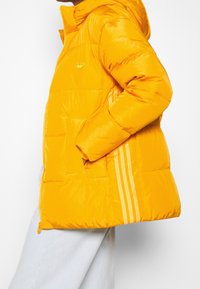 Gul puffjacka med en glänsande textur, som har en zippered framsida, fickor och tre vita ränder längs sidan. Adidas-logotyp på bröstet.