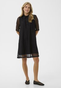 Sieviete ģērbta ceļgala garuma melnā mežģīņu kleitā ar apkakli un pusmēteļiem, apavi ir melnas austas plakanas kurpes, stāvot uz balta fona.