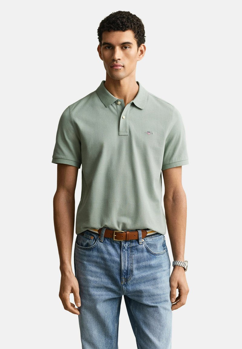 Junger Mann, der ein hellgrünes Poloshirt, blaue Jeans, einen braunen Gürtel und eine silberne Armbanduhr trägt, vor einem weißen Hintergrund stehend.