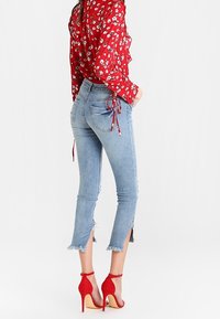Jeans de mezclilla azul claro de cintura alta con dobladillo deshilachado y aberturas laterales, combinados con una camisa roja de flores con botones y zapatos de tacón rojos.