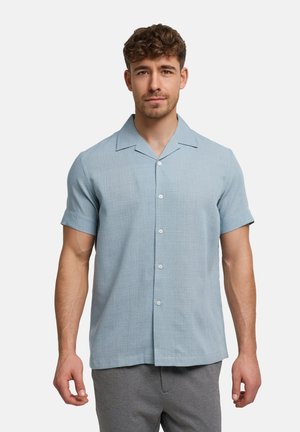 Man staat met het gezicht naar voren, draagt een lichtblauw overhemd met korte mouwen en een grijze broek tegen een effen witte achtergrond.