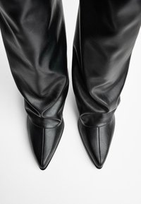 Des bottes noires en cuir à bout pointu présentent une texture lisse et un design ajusté, avec un léger drapage au niveau de la cheville pour une silhouette élégante.