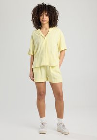 Camicia a maniche corte giallo a righe con bottoni abbinata a pantaloni corti della stessa fantasia. Il tessuto sembra leggero e ha una consistenza morbida. Indossa sneakers bianche.