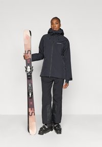 Sort skijakke med hætte og lynlåslommer, sorte skibukser og sorte skistøvler. Holder i pink og sorte ski med bindinger.