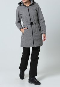 Manteau gris à manches longues avec taille cintrée, fermeture éclair et capuche. Associé à un pantalon noir et des bottes de randonnée noires.
