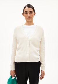 Cremefarbener Cardigan mit V-Ausschnitt, Knopfverschluss, gerippten Bündchen und Saum. Über einem weißen Hemd getragen, von einer Person in schwarzen Hosen gehalten.