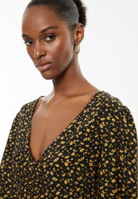 Zwart bloemenjurk met gele accenten, V-hals ontwerp en puffmouwen. Model draagt gouden hoopoorbellen. Natuurlijk haar gestyled in een knot.