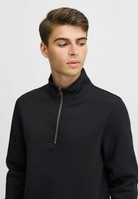 Pullover en polaire noir avec fermeture éclair, col haut, texture lisse, sans motifs ni accents supplémentaires.