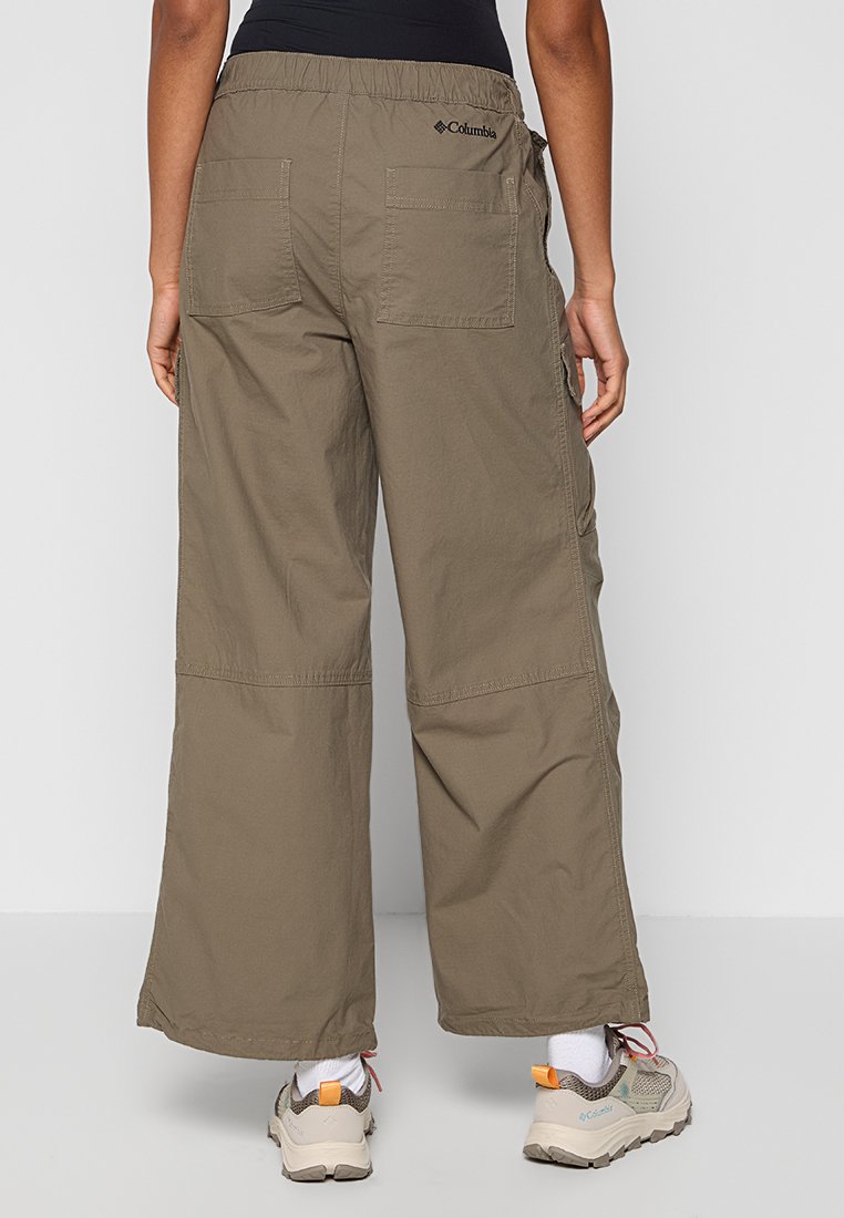 Osoba ubrana w luźne oliwkowe spodnie cargo z tylnymi kieszeniami oraz buty trekkingowe z pomarańczowymi uchwytami, stojąca na jednolitym tle.
