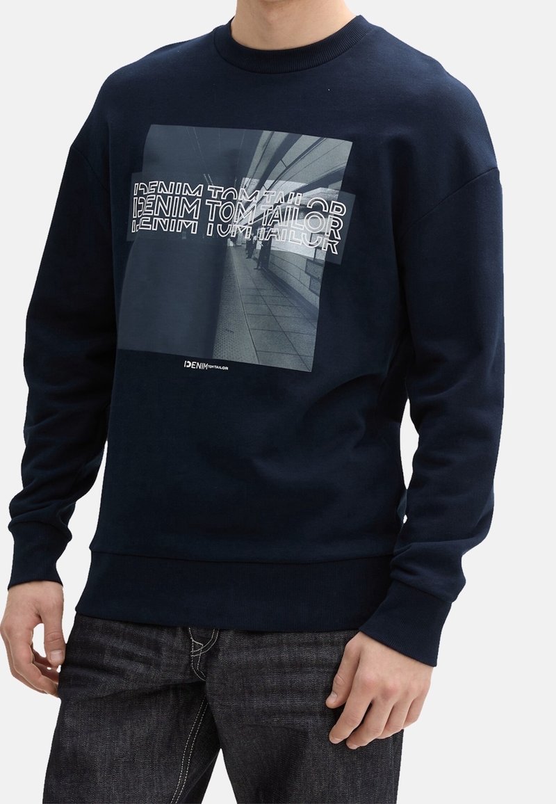 Homme portant un sweat-shirt bleu marine avec un motif graphique en noir et blanc d'un couloir urbain et le texte superposé "DENIM TOM TAILOR" sur le devant.
