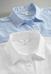 Due camicie a collar, sovrapposte; una azzurro chiaro con strisce bianche, l'altra bianca semplice. Entrambe le camicie presentano una chiusura con bottoni davanti e un'etichetta.