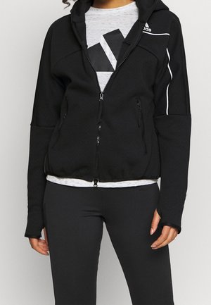 Kobieta nosząca czarną bluzę z kapturem zapinaną na zamek, z białymi lamówkami i logo Adidas, na szarej koszulce z dużą czarną grafiką, w połączeniu z czarnymi legginsami.
