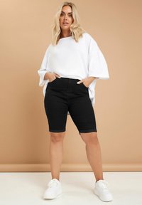 Camiseta blanca oversized, pantalones cortos de mezclilla negros con dobladillos enrollados, combinados con zapatillas blancas. Colores sólidos y corte relajado.