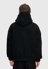 Persona con capelli ricci che indossa una giacca in pile nera con cappuccio, vista da dietro su uno sfondo grigio chiaro uniforme.