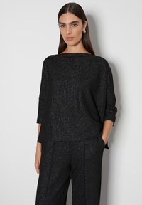 Schwarzes Oberteil mit Dreiviertelärmeln, das aus einem strukturierten Stoff besteht und mit dezenten Glitzerakzenten versehen ist, passend zu maßgeschneiderten schwarzen Hosen.